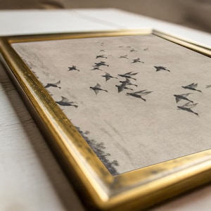 May include: A framed print featuring a flock of birds in flight against a neutral background. The birds are depicted in shades of gray, creating a sense of movement. The artwork is encased in a gold-colored frame, adding a touch of elegance.