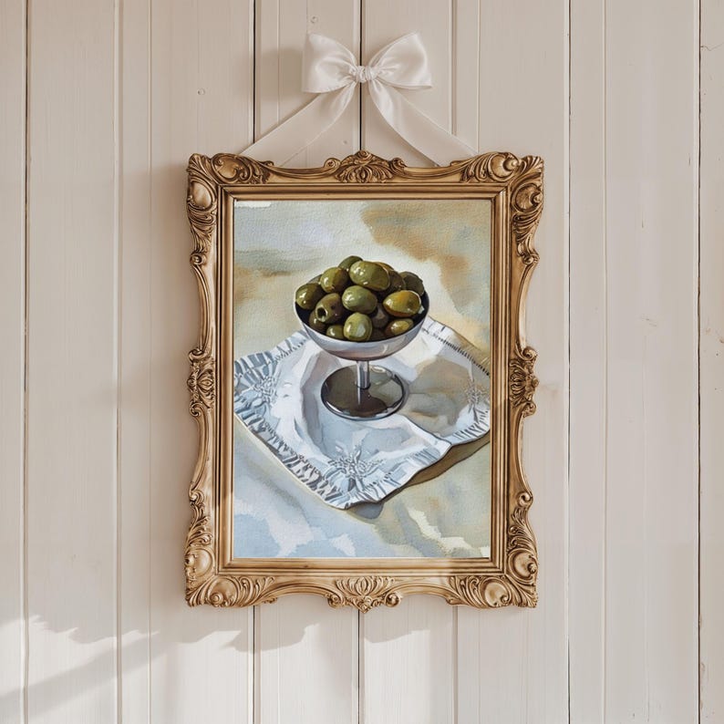 May include: A framed painting of green olives in a silver bowl on a white cloth. The painting is in a gold ornate frame and is hanging on a white wall. A white bow is tied at the top.