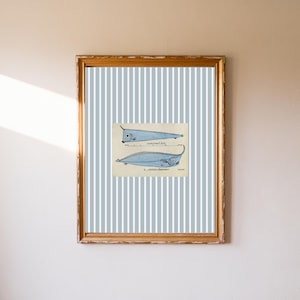 Puede incluir: Impresión enmarcada que presenta dos ilustraciones estilizadas de peces, cada una con un cuerpo largo y delgado y un solo ojo. Los peces están representados en tonos de azul y gris sobre un fondo beige. La impresión está colocada sobre un fondo a rayas y enmarcada en un marco dorado.
