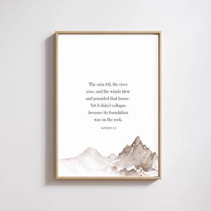 May include: A framed print with a light wood-coloured frame. The print features a watercolour mountain scene in brown and grey tones, and text from Matthew 7:25. The text is in a simple, elegant font.