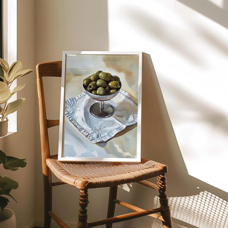 May include: A framed artwork depicting a still life of green olives in a silver bowl. The bowl sits on a white cloth with a decorative border. The artwork is displayed on a wooden chair, with sunlight casting shadows.
