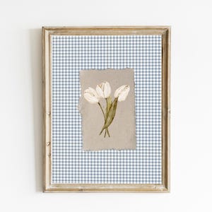 Op de afbeelding: Ingelijst kunstwerk met een aquarel illustratie van drie witte tulpen. Het kunstwerk is geplaatst tegen een blauw-wit ruitpatroon, omlijst in een goudkleurige houten lijst. Het is een bloemenprint.