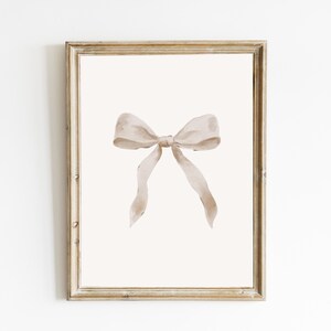 May include: A framed print showing a watercolour illustration of a beige bow. The bow is centrally placed on a cream background, within a gold-toned wooden frame. The artwork has a soft, elegant aesthetic.