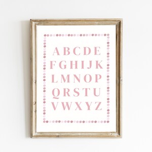 May include: A framed alphabet print featuring the letters A through Z in a soft pink hue. The letters are centered on a white background, surrounded by a dotted border in varying shades of pink. The frame is a light, natural wood color.