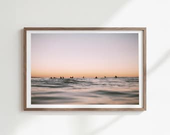 May include: A framed print depicting a tranquil ocean scene at sunset. Silhouetted figures of surfers are seen on the water's surface. The sky transitions from a soft pink to a warm orange hue, creating a peaceful atmosphere.