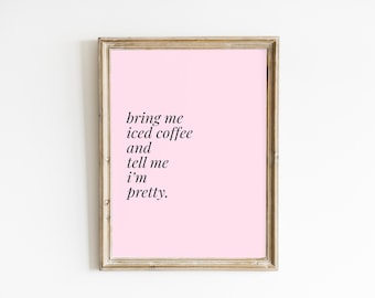 May include: A framed print with a pink background and black text that reads, "bring me iced coffee and tell me I'm pretty." The frame is a light brown colour. The print is displayed against a white wall.