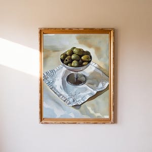 May include: A framed painting depicts a silver goblet filled with green olives, resting on a white, patterned cloth. The painting has a gold-colored frame and is set against a neutral background. The artwork captures a still life scene.