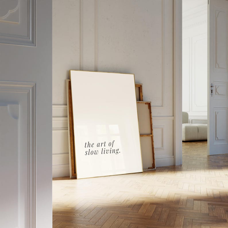 May include: A large, white canvas with the text "the art of slow living" leans against a wall. The canvas has a wooden frame. The image is taken in a room with a hardwood floor and a doorway leading to another room.