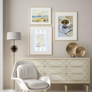May include: Three framed nautical-themed watercolor paintings hang on a neutral wall. One depicts a sailboat on the sea, another shows olives in a glass bowl, and the third features a sailboat with the text "Sailing Club". A white chair and a gold floor lamp are in the foreground.