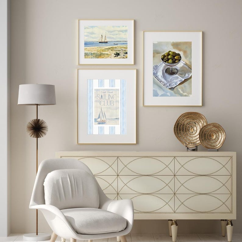 May include: Three framed nautical-themed watercolor paintings hang on a neutral wall. One depicts a sailboat on the sea, another shows olives in a glass bowl, and the third features a sailboat with the text "Sailing Club". A white chair and a gold floor lamp are in the foreground.