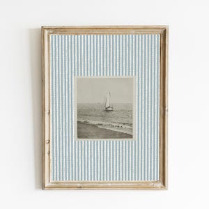 May include: Framed nautical art print featuring a sailboat on the ocean. The artwork is set within a light blue and white striped border, encased in a wooden frame. The overall aesthetic is vintage and coastal.