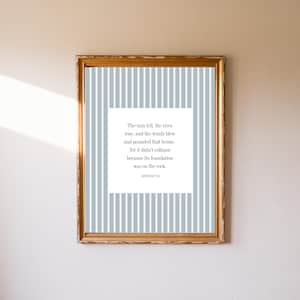 May include: A framed print with a gold-colored frame. The print features a white square with text on a light blue and white striped background. The text reads: "The rain fell, the river rose, and the winds blew and pounded that house. Yet it didn't collapse because its foundation was on the rock."