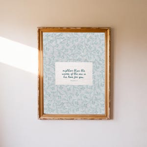 Puede incluir: Una impresi&oacute;n enmarcada con un marco dorado. La impresi&oacute;n presenta un fondo floral azul claro y un espacio rectangular blanco con el texto "mightier than the waves of the sea is his love for you."
