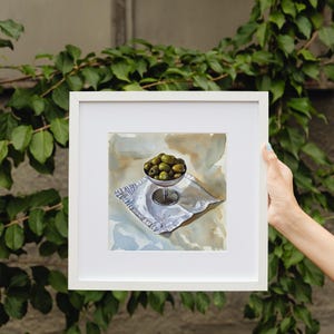 May include: A framed watercolor painting of a still life. The painting depicts a small silver bowl filled with green olives, resting on a white, patterned napkin. The background has neutral tones. The frame is white.