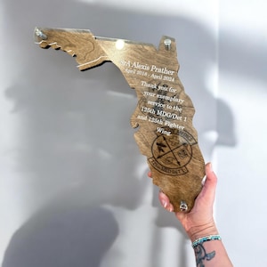 May include: A wooden plaque in the shape of Florida, held by a hand. The plaque has engraved text: "SrA Alexis Prather April 2018 - April 2024" and a military emblem. The wood is a warm brown color.