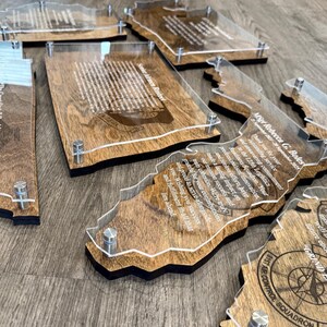 May include: A collection of wooden plaques with clear acrylic overlays, each shaped like a state or region. The plaques feature engraved text and are held together with silver-colored hardware. The wood has a warm, natural tone.