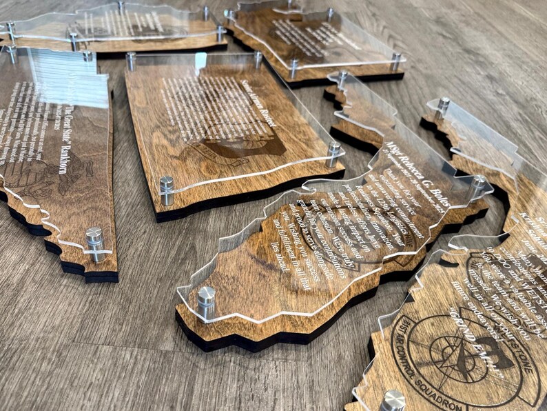 May include: A collection of wooden plaques with clear acrylic overlays, each shaped like a state or region. The plaques feature engraved text and are held together with silver-colored hardware. The wood has a warm, natural tone.