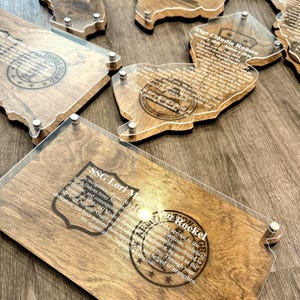 May include: Wooden plaques with clear acrylic overlays, featuring engraved text and state-shaped designs. The plaques have a natural wood grain finish and are secured with silver-colored hardware. The text includes names and military-related phrases.