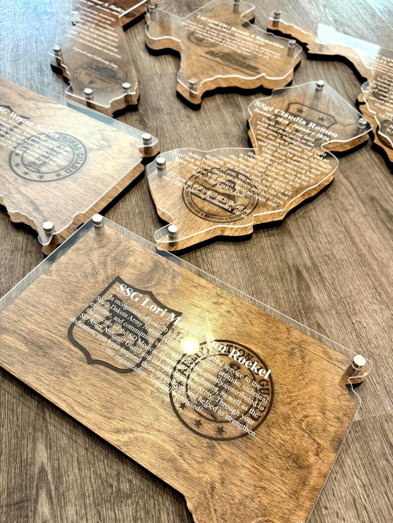 May include: Wooden plaques with clear acrylic overlays, featuring engraved text and state-shaped designs. The plaques have a natural wood grain finish and are secured with silver-colored hardware. The text includes names and military-related phrases.