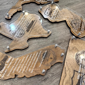 May include: A collection of wooden plaques in various state shapes, each with a clear acrylic overlay. The plaques feature engraved text and designs, including names, dates, and military insignia. The wood has a warm, natural tone, and the acrylic is held in place with silver hardware.