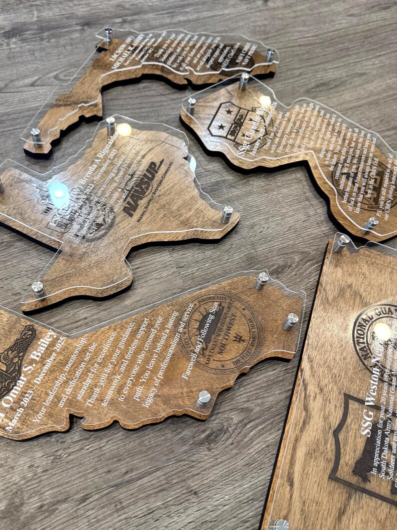 May include: A collection of wooden plaques in various state shapes, each with a clear acrylic overlay. The plaques feature engraved text and designs, including names, dates, and military insignia. The wood has a warm, natural tone, and the acrylic is held in place with silver hardware.