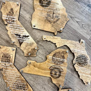 May include: Wooden state-shaped plaques with clear acrylic overlays. Each plaque features engraved text and military insignia, including the states of California, Georgia, Florida, and New York. The plaques are fastened with silver screws.