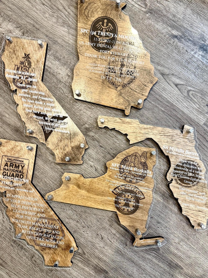 May include: Wooden state-shaped plaques with clear acrylic overlays. Each plaque features engraved text and military insignia, including the states of California, Georgia, Florida, and New York. The plaques are fastened with silver screws.