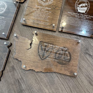 May include: A collection of wooden plaques with clear acrylic overlays, featuring engraved text and military emblems. The plaques are in various shapes, including a state outline, and are mounted with silver-colored hardware.