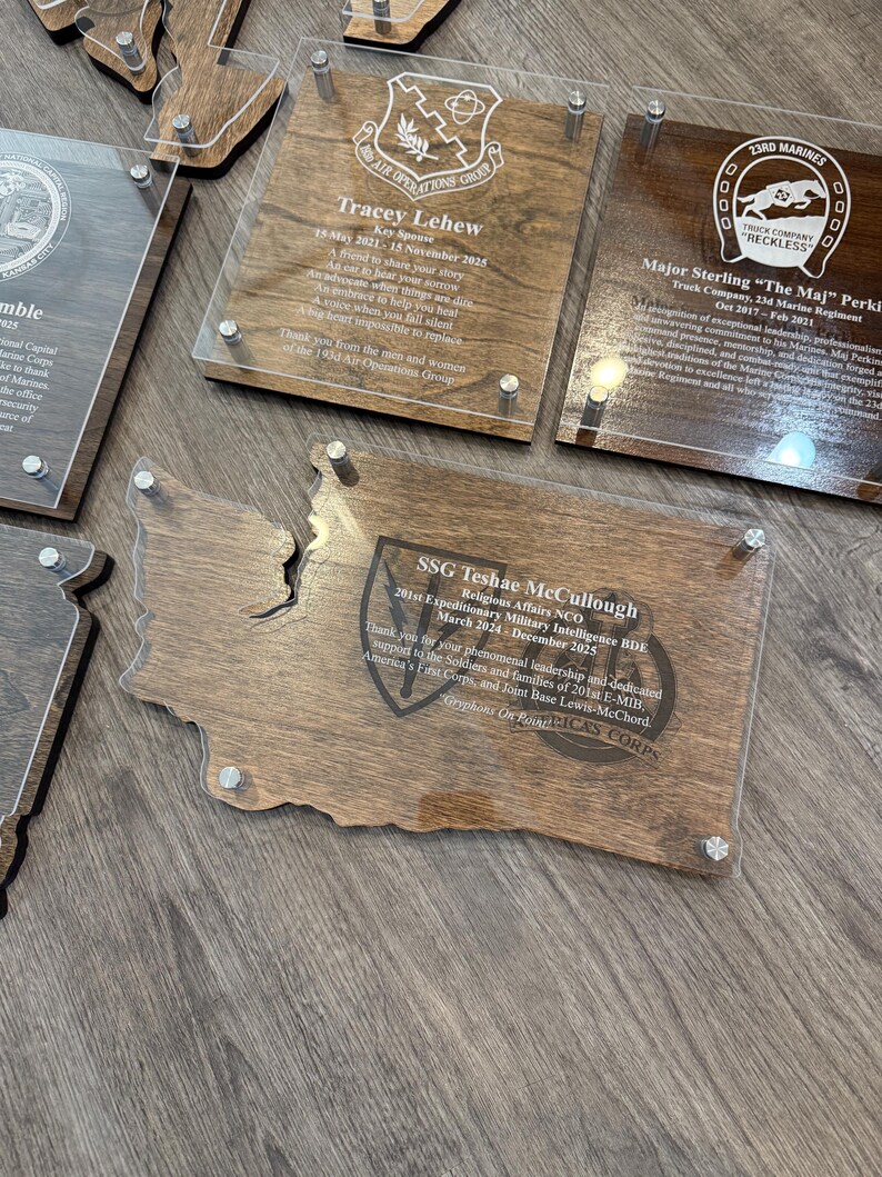 May include: A collection of wooden plaques with clear acrylic overlays, featuring engraved text and military emblems. The plaques are in various shapes, including a state outline, and are mounted with silver-colored hardware.