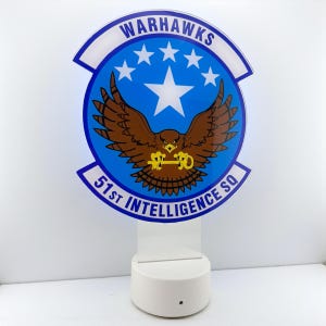 Op de afbeelding: Een verlicht bord met het Warhawks-embleem, een bruine adelaar met een sleutel, tegen een blauwe achtergrond met witte sterren. De tekst "WARHAWKS" en "51st INTELLIGENCE SQ" zijn zichtbaar. De basis is wit.