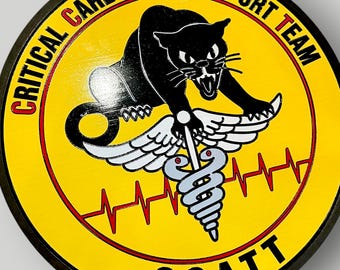 May include: A circular patch featuring a yellow background with a black panther graphic holding a white caduceus. The text reads CRITICAL CARE AIR TRANSPORT TEAM and CCATT. A red EKG line runs horizontally across the center of the yellow field.