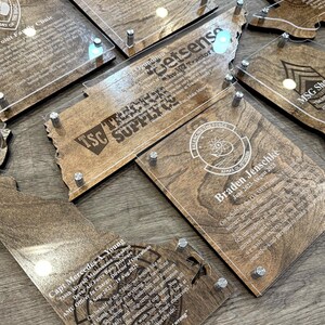 May include: A collection of wooden plaques with clear acrylic overlays, featuring engraved text and designs. The plaques are in various shapes, including state outlines, and are secured with silver-colored hardware. The text includes names, dates, and organizational logos.