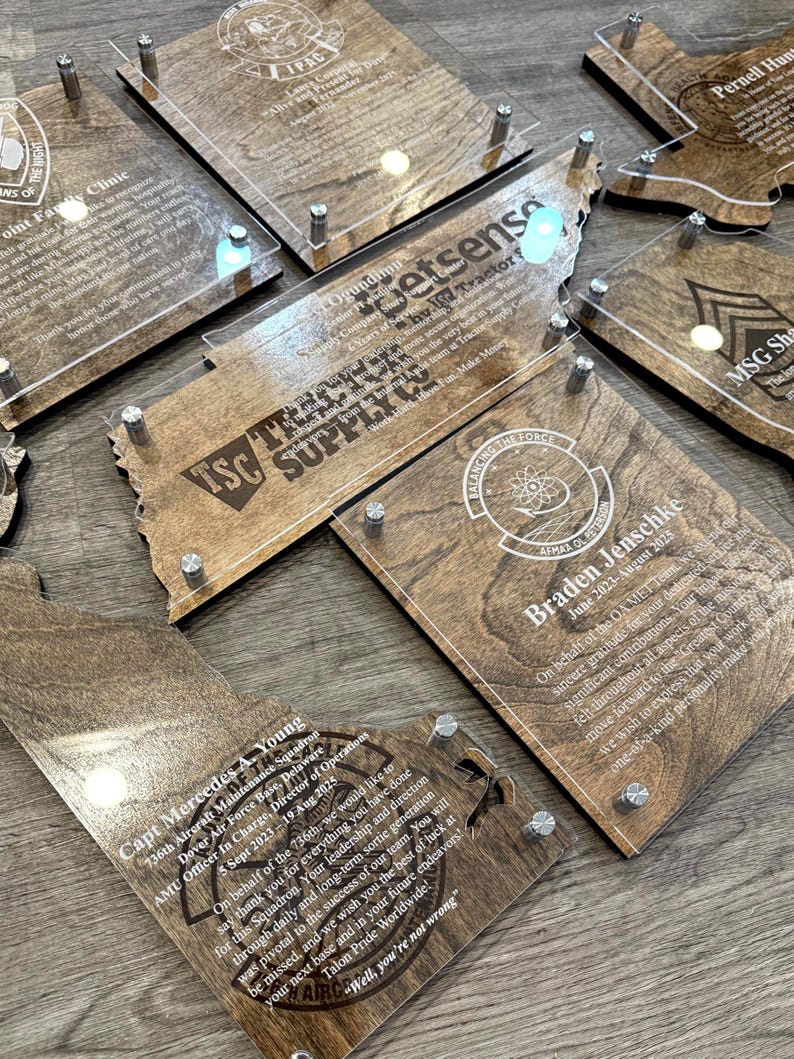 May include: A collection of wooden plaques with clear acrylic overlays, featuring engraved text and designs. The plaques are in various shapes, including state outlines, and are secured with silver-colored hardware. The text includes names, dates, and organizational logos.