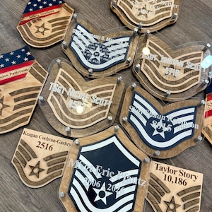 May include: A collection of custom wooden and acrylic military rank plaques featuring engraved names, dates, and service insignias. Each piece displays layered wood textures, clear acrylic overlays, and metallic hardware, showcasing various Air Force rank designs.