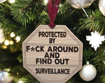 May include: A wooden Christmas ornament shaped like a stop sign with the text "Protected by F*ck Around and Find Out Surveillance" in black lettering.