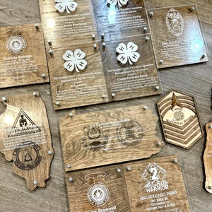 May include: A collection of wooden and clear acrylic awards and plaques. The plaques feature engraved text and logos, including 4-H clover designs, military emblems, and names. The awards are fastened with silver-colored hardware.