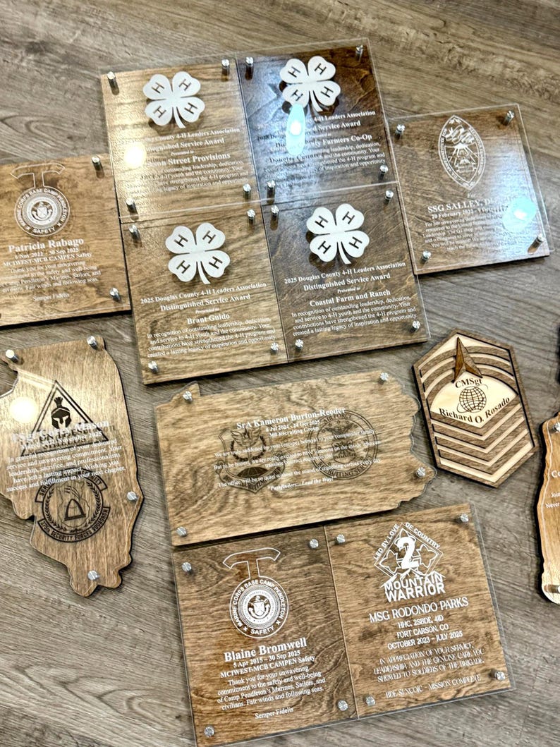 May include: A collection of wooden and clear acrylic awards and plaques. The plaques feature engraved text and logos, including 4-H clover designs, military emblems, and names. The awards are fastened with silver-colored hardware.