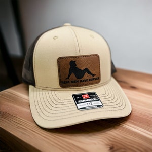 May include: A beige and brown trucker hat with a brown leather patch that says "Real Men Have Curves". The hat has a black silhouette of a person sitting down on the patch. The hat also has a red and black Richardson 112 label.