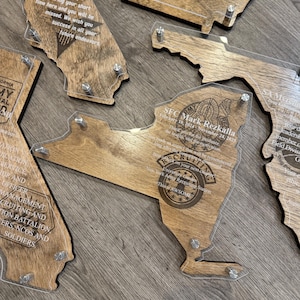 May include: Wooden plaques in the shape of U.S. states, with clear acrylic overlays. The plaques feature engraved text and are held together with silver-colored hardware. The plaques are a light brown color.