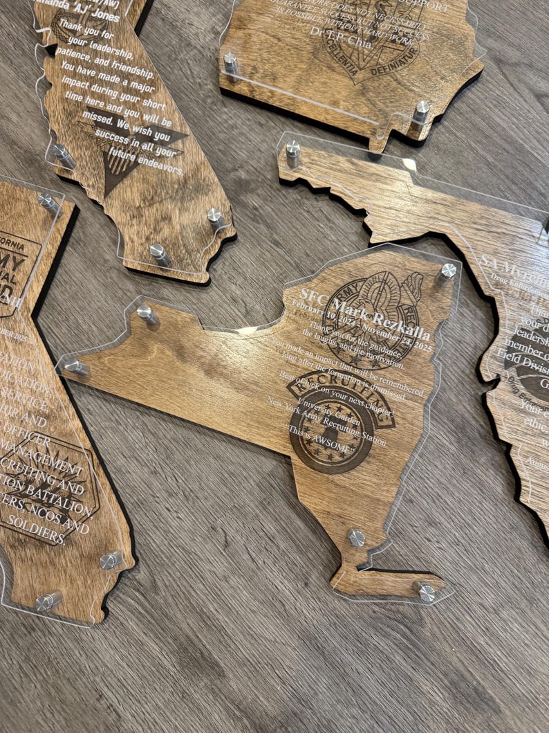 May include: Wooden plaques in the shape of U.S. states, with clear acrylic overlays. The plaques feature engraved text and are held together with silver-colored hardware. The plaques are a light brown color.