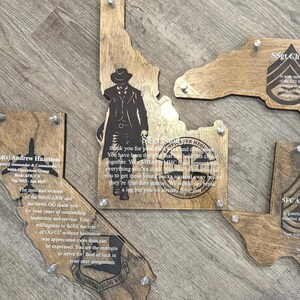 May include: Wooden plaques in the shape of U.S. states, with clear acrylic overlays. The plaques feature engraved text and images, including a cowboy silhouette on the Idaho plaque. The plaques are mounted with silver hardware.