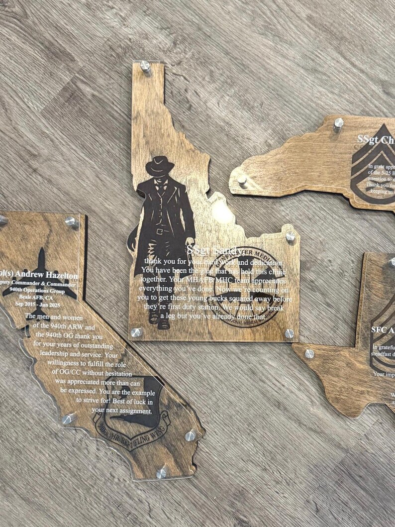 May include: Wooden plaques in the shape of U.S. states, with clear acrylic overlays. The plaques feature engraved text and images, including a cowboy silhouette on the Idaho plaque. The plaques are mounted with silver hardware.