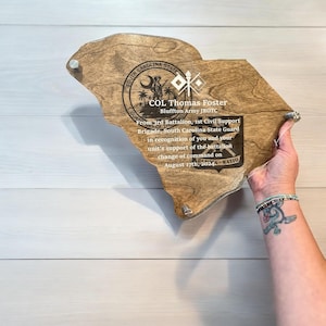 May include: A wooden plaque in the shape of South Carolina, with text that reads "COL Thomas Foster" and "August 17th, 2024". The plaque has a clear acrylic front and is held by a person.