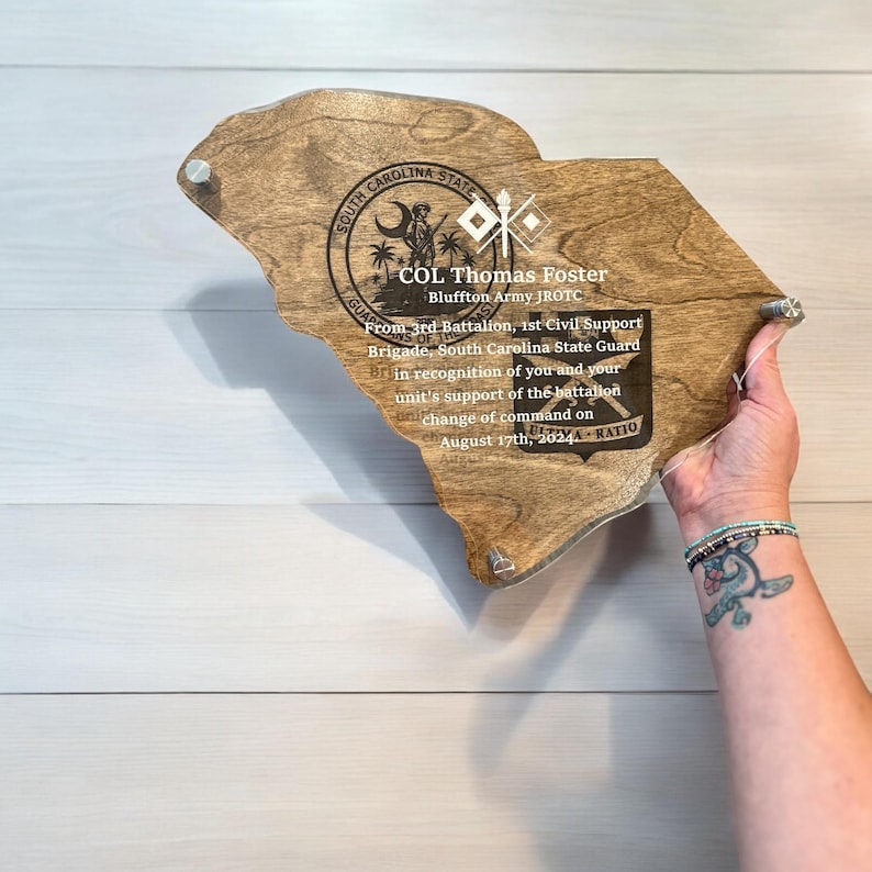 May include: A wooden plaque in the shape of South Carolina, with text that reads "COL Thomas Foster" and "August 17th, 2024". The plaque has a clear acrylic front and is held by a person.