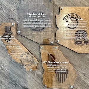 May include: Wooden plaques in the shape of U.S. states, engraved with text and military emblems. The plaques are mounted on a wooden surface and secured with silver-colored hardware. The plaques are for military personnel.