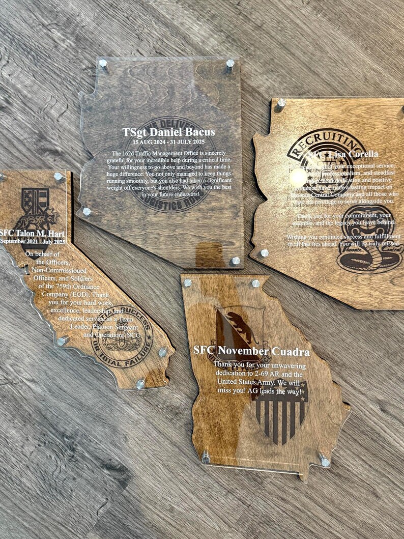 May include: Wooden plaques in the shape of U.S. states, engraved with text and military emblems. The plaques are mounted on a wooden surface and secured with silver-colored hardware. The plaques are for military personnel.