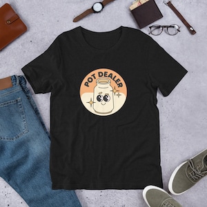 Puede incluir: Camiseta negra con una ilustración de dibujos animados de un frasco con ojos y el texto "POT DEALER" en un círculo.