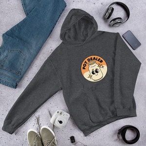 May include: A dark gray hooded sweatshirt with a cartoon illustration of a can with a face and the text "POT DEALER" in a circle.