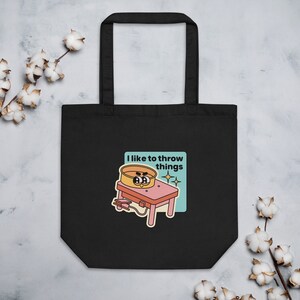 May include: Black tote bag with a white design of a pottery wheel with a face and the text "I like to throw things".