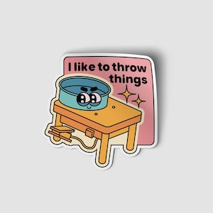 May include: A sticker with a cartoon illustration of a pottery wheel with a face. The wheel is on a wooden table with a foot pedal. The sticker has the text "I like to throw things" in black text on a pink background.